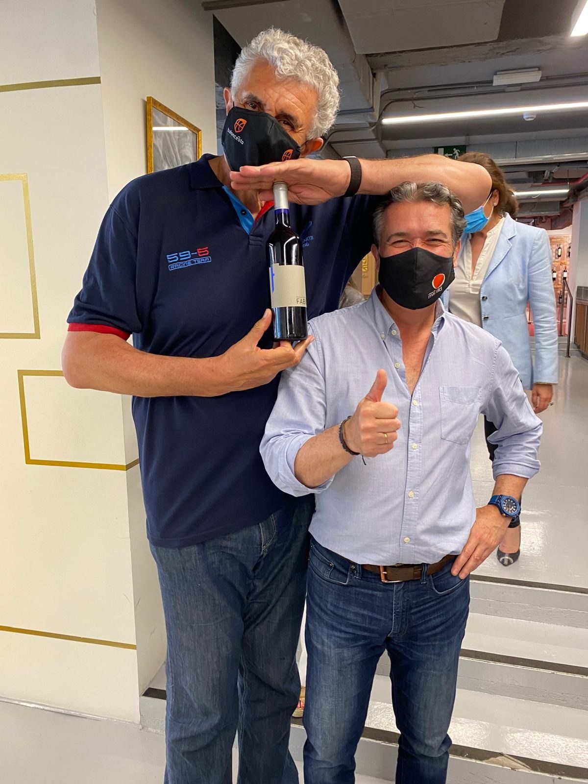 Dos hombres con máscaras faciales están de pie uno al lado del otro sosteniendo una botella de vino.