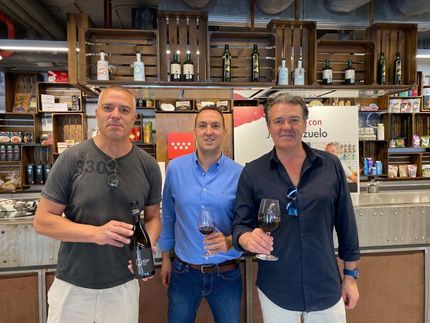 Tres hombres están de pie uno al lado del otro sosteniendo copas de vino y botellas de vino.
