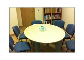 Pequeña sala de reuniones con una mesa redonda, cuatro sillas azules y una estantería al fondo.