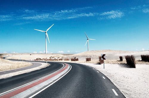 Carretera sinuosa por el desierto junto a dunas de arena blanca y aerogeneradores bajo un cielo azul despejado