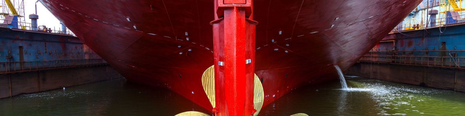 Casco rojo de un barco en un dique, centrado sobre una quilla roja, sobre hormigón y con agua a ambos lados.