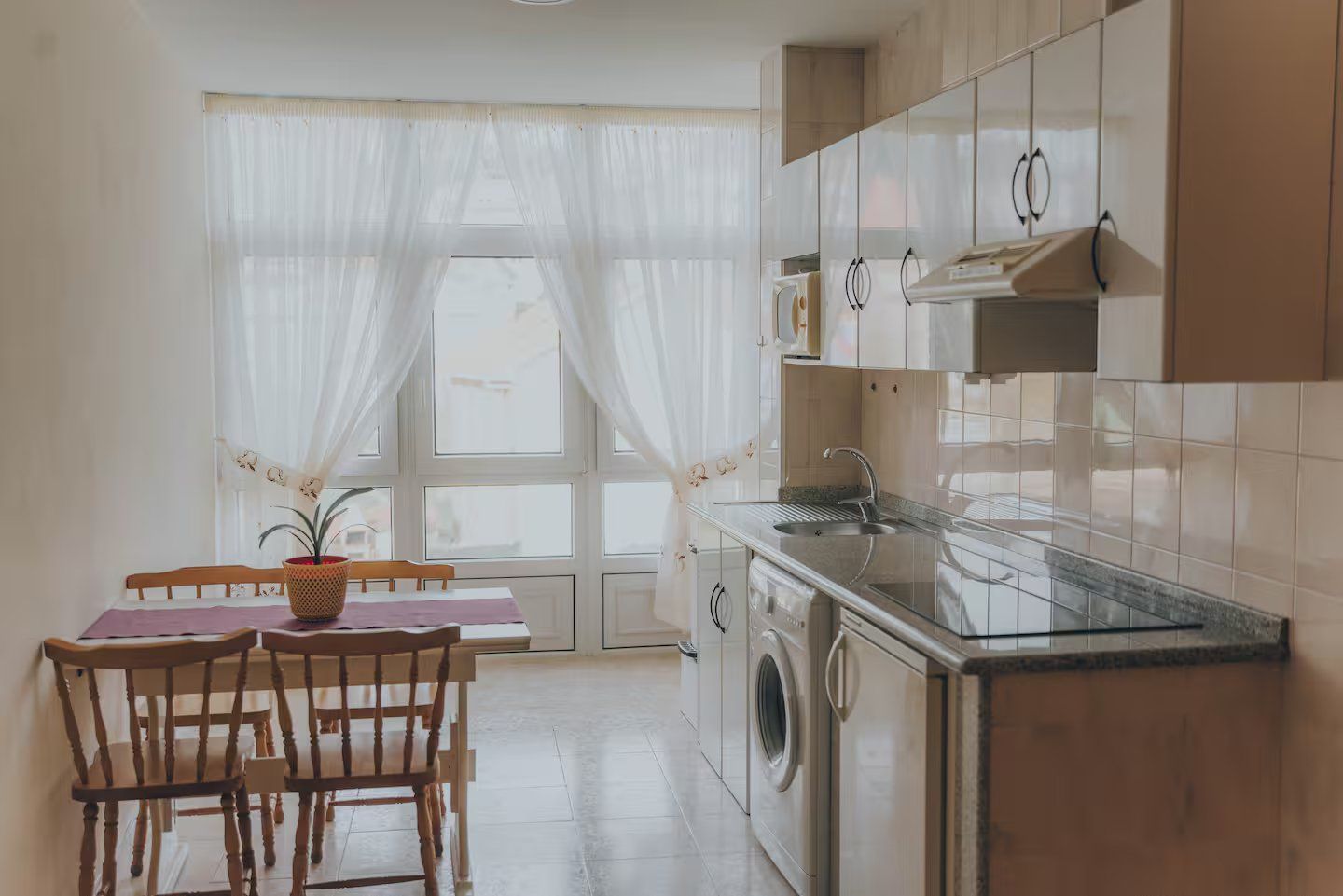 Una cocina con armarios blancos, una encimera con vitrocerámica y lavadora, y una pequeña mesa de madera con sillas.