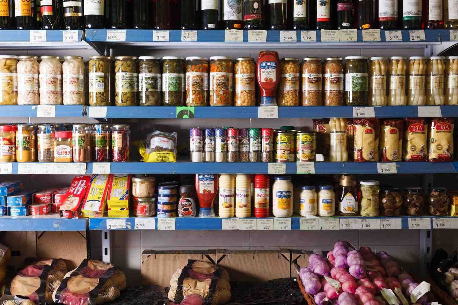Estantes de un supermercado repletos de frascos surtidos, condimentos, productos envasados, patatas y cebollas moradas.