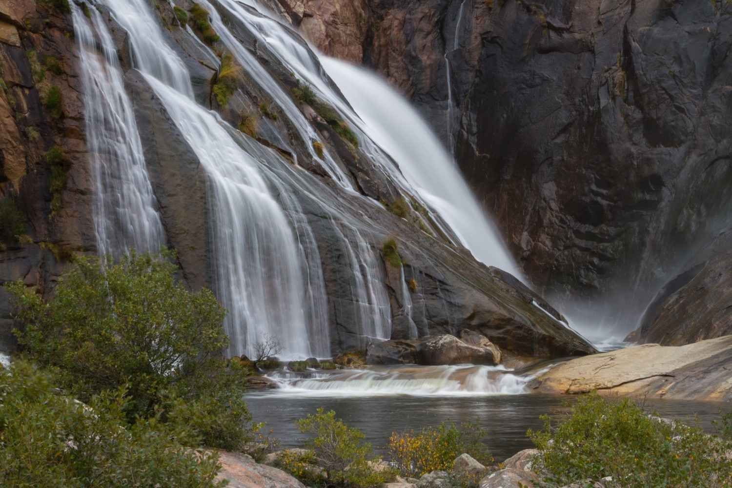 Una cascada cae en cascada sobre lisas paredes de roca oscura hasta una tranquila poza, rodeada de vegetación en su base.