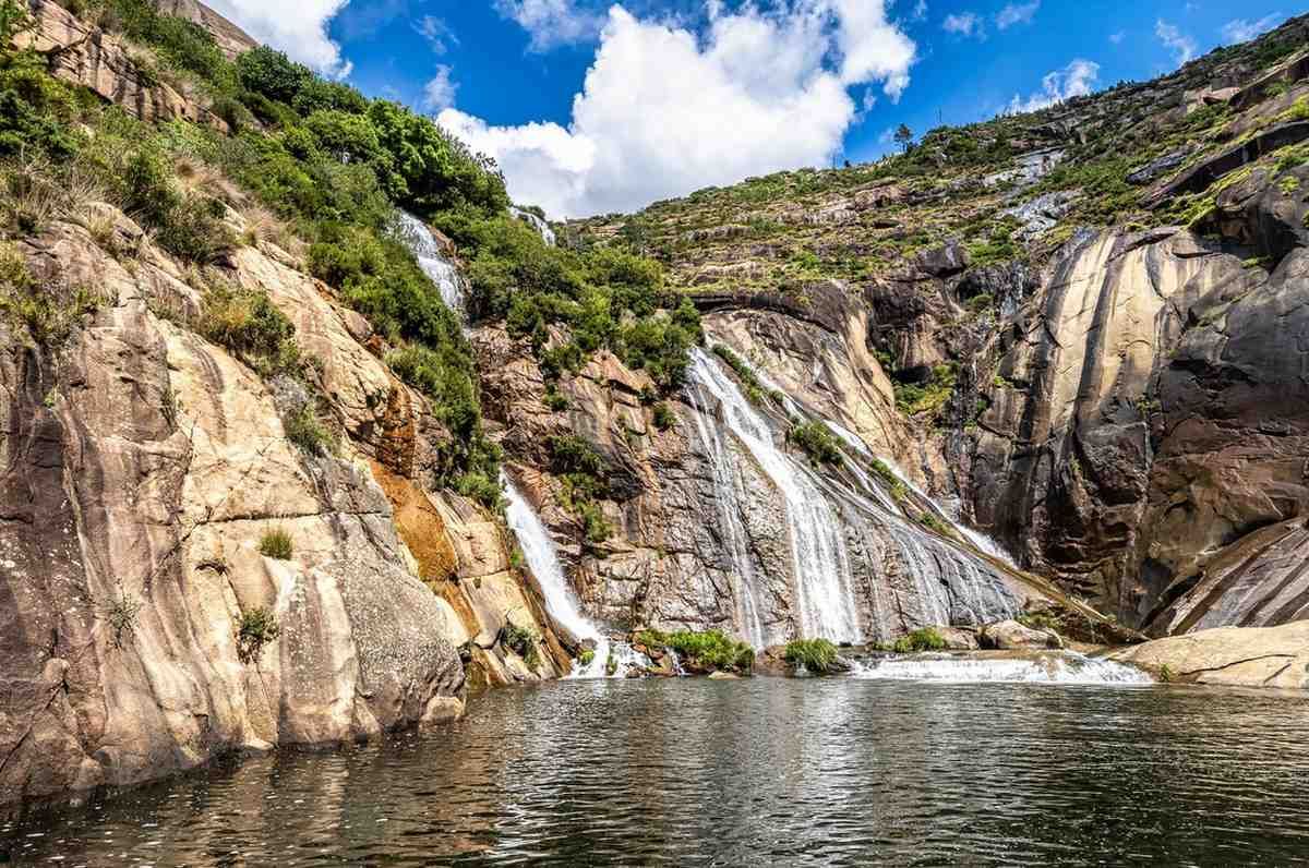 Una cascada cae en picado por escarpados acantilados rocosos hasta formar una poza bajo un cielo azul brillante y nublado.
