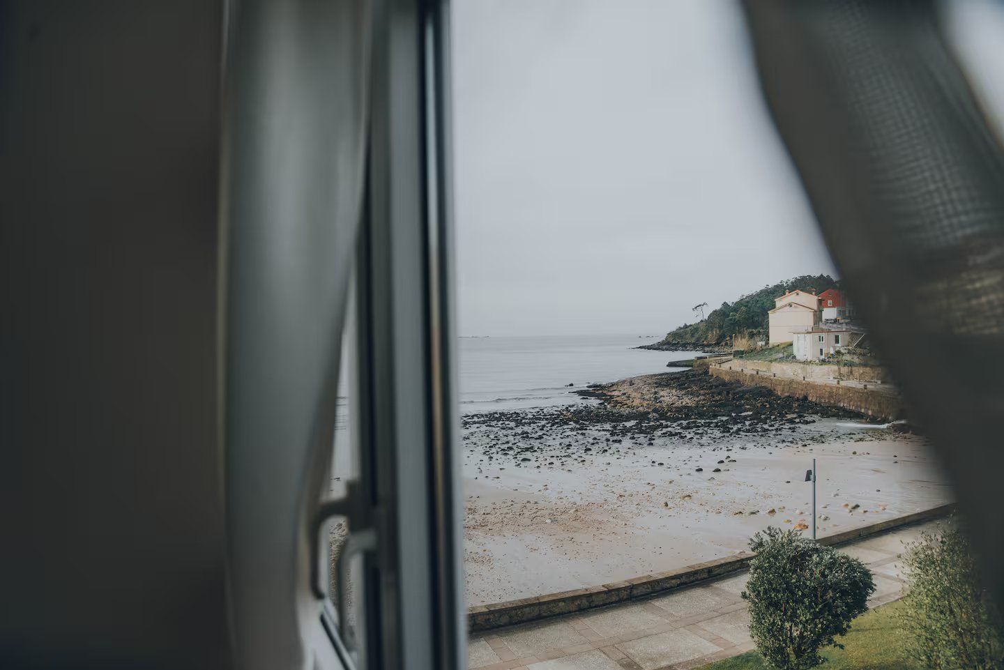 Una vista a través de una ventana que muestra una playa rocosa, aguas tranquilas y un edificio en la costa.