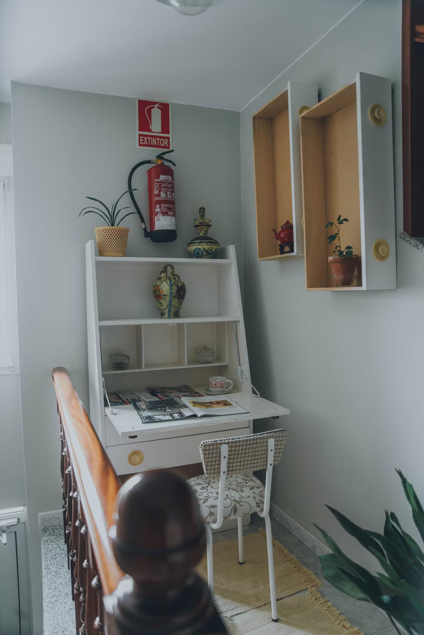 Un pequeño escritorio blanco con una silla y elementos decorativos contra una pared gris, con un extintor.