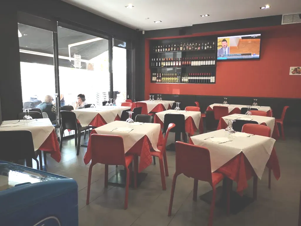 Interior de restaurante con mesas, sillas rojas y una pared roja. La gente está sentada frente a los grandes ventanales.