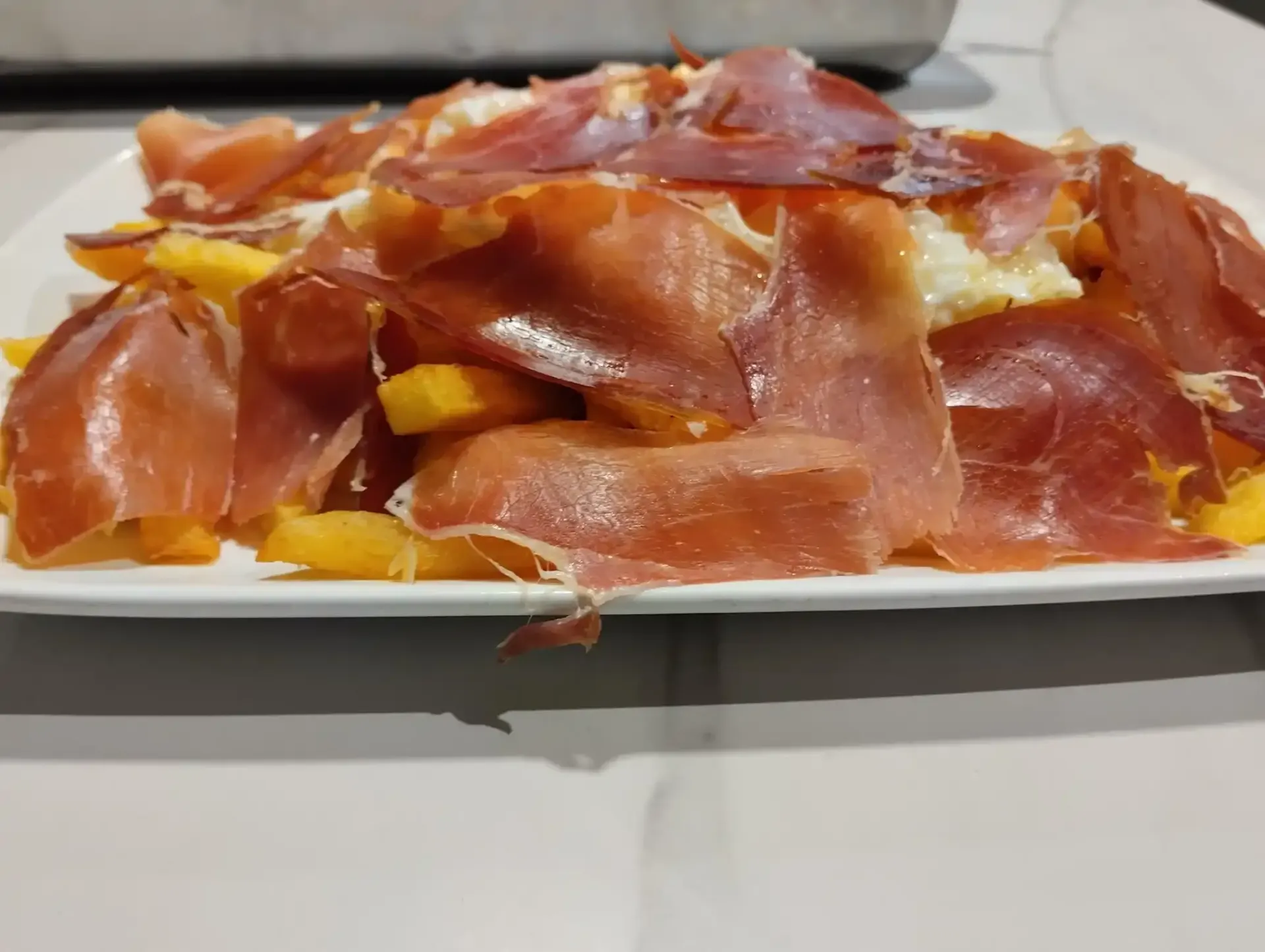 Plato de comida: patatas amarillas cortadas en cubos cubiertas con finas lonchas de jamón curado.