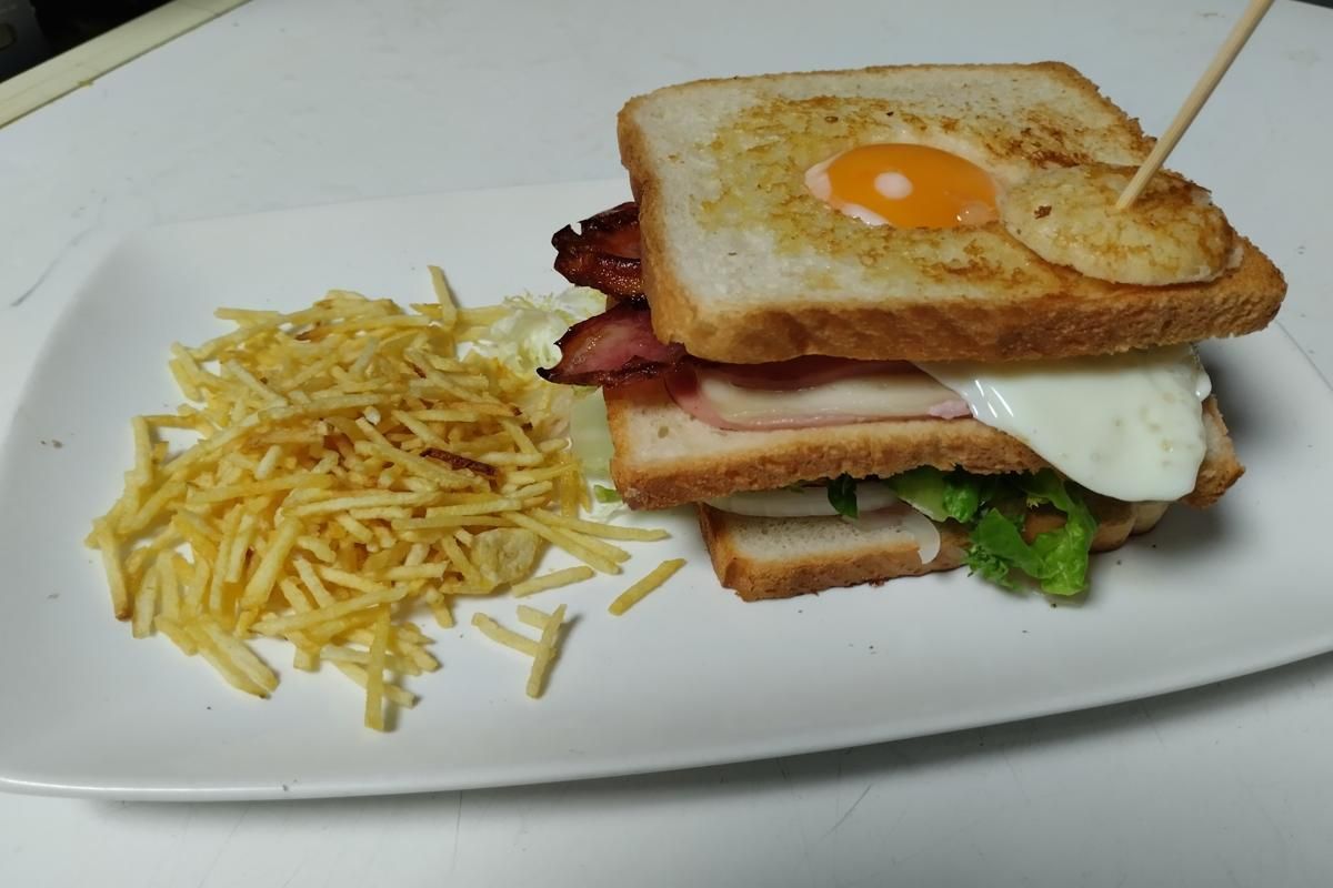 Sándwich club con huevo frito, tocino, jamón, lechuga y papas fritas en un plato blanco.