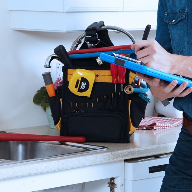 Un plombier écrit sur un bloc-notes à côté d'une sacoche à outils remplie d'outils, posée sur un comptoir de cuisine près d'un évier.
