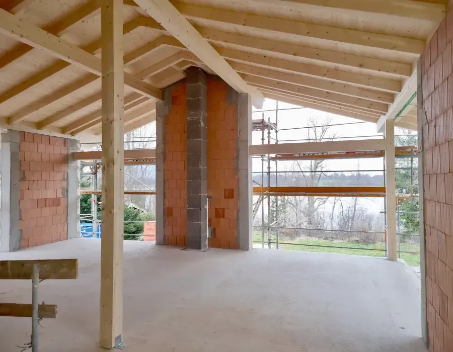 Innenansicht eines Neubaus im Rohbau mit sichtbarer Holzdachkonstruktion und Mauerwerk.