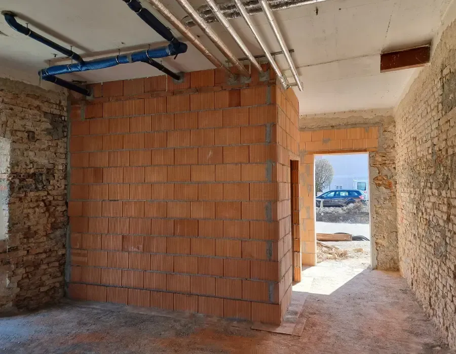 Innenansicht eines Anbaus im Rohbau mit neu errichteten Ziegelwänden und sichtbaren Installationsleitungen an der Decke.