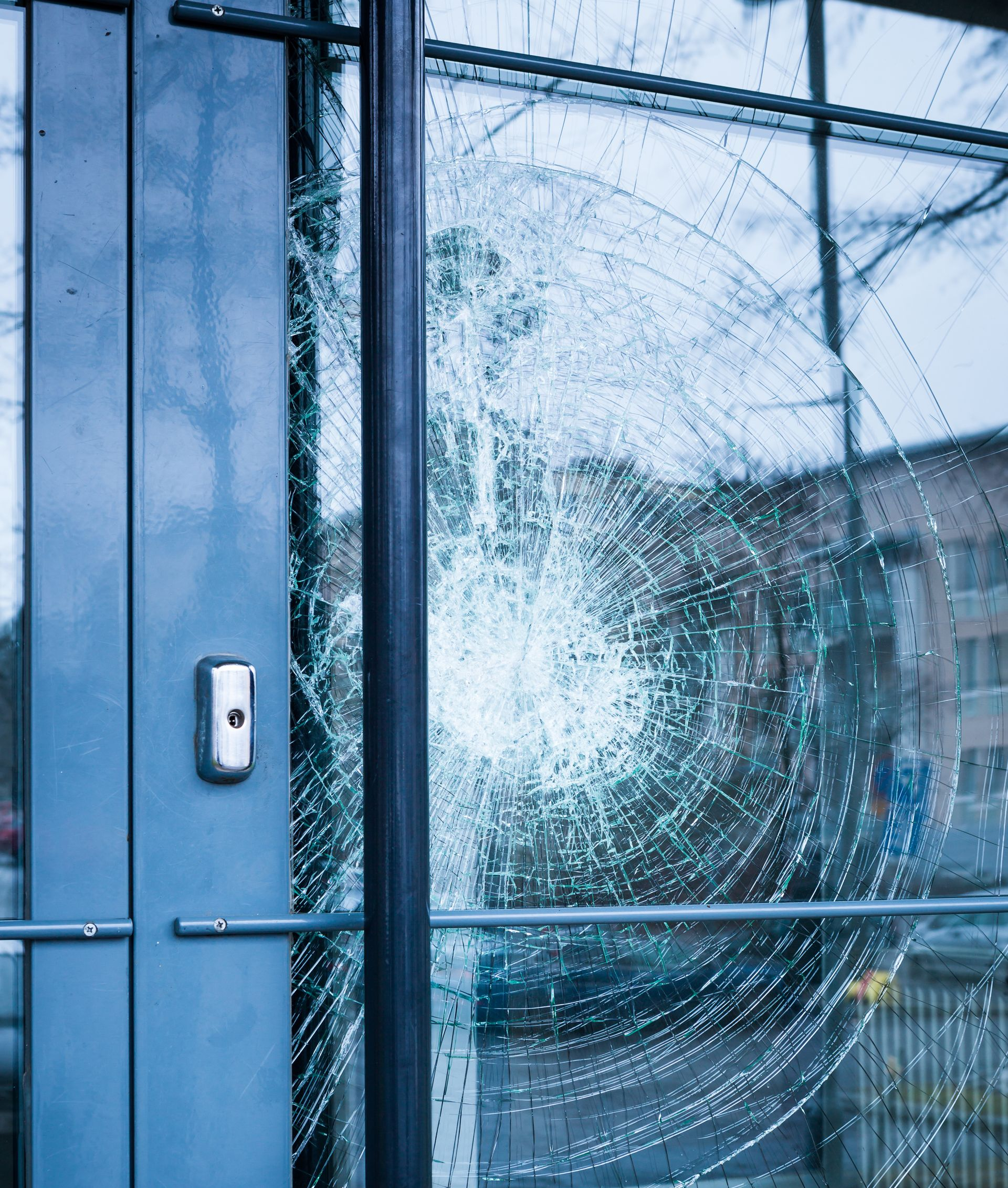 Porte vitrée brisée ; éclats de verre disposés en cercle, quincaillerie de porte visible et arrière-plan du bâtiment.