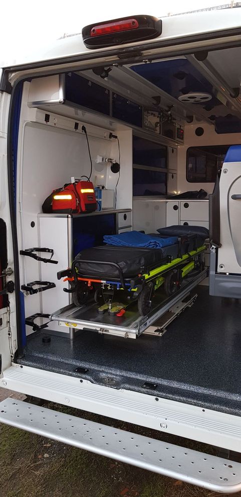 La porte arrière ouverte d'une ambulance révèle un brancard vide sur un rail métallique à l'intérieur du compartiment patient aux parois.