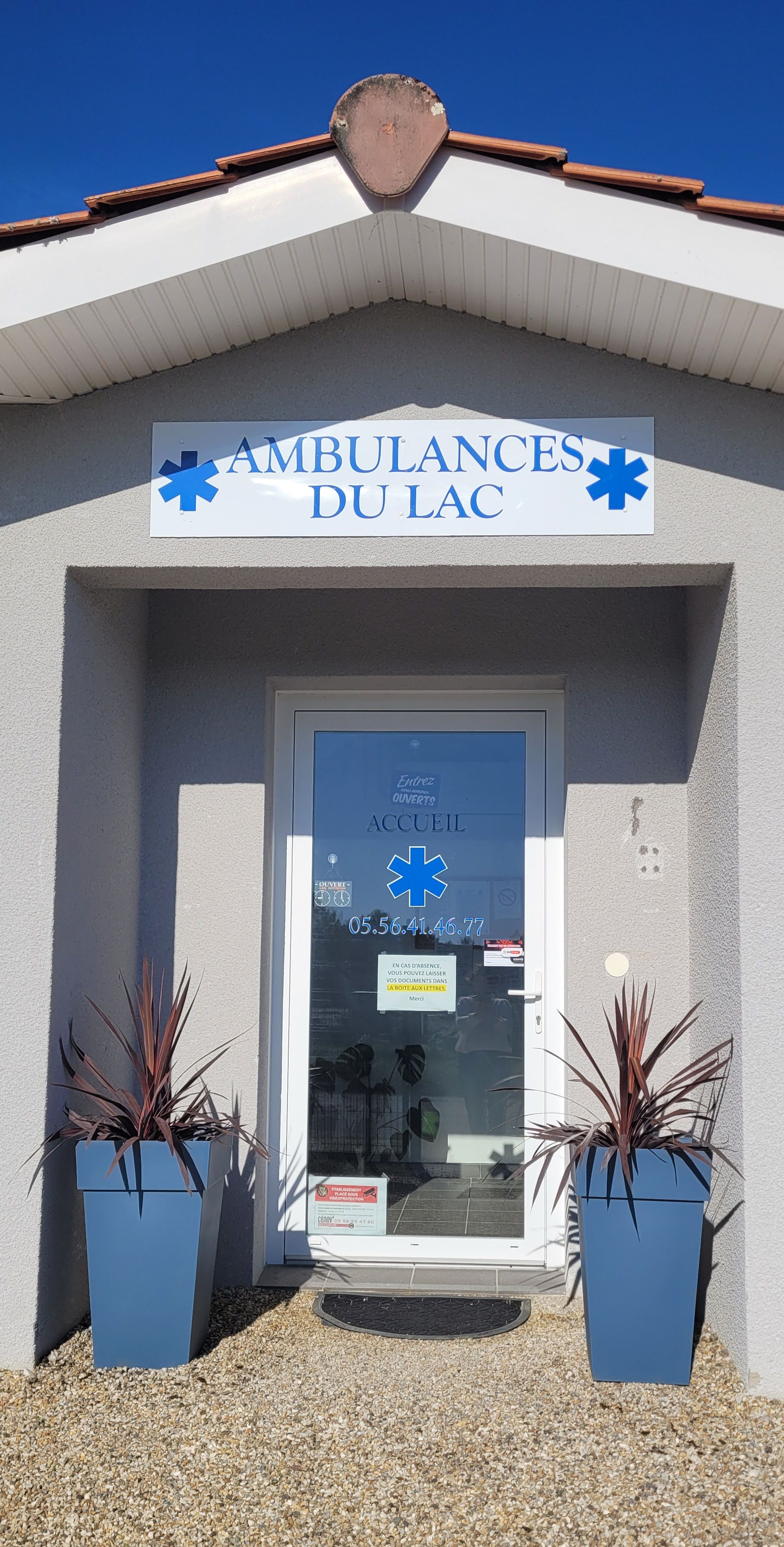 Entrée d'Ambulances du Lac avec un panneau, le logo de l'étoile de vie et des plantes en pot sur un chemin de gravier.