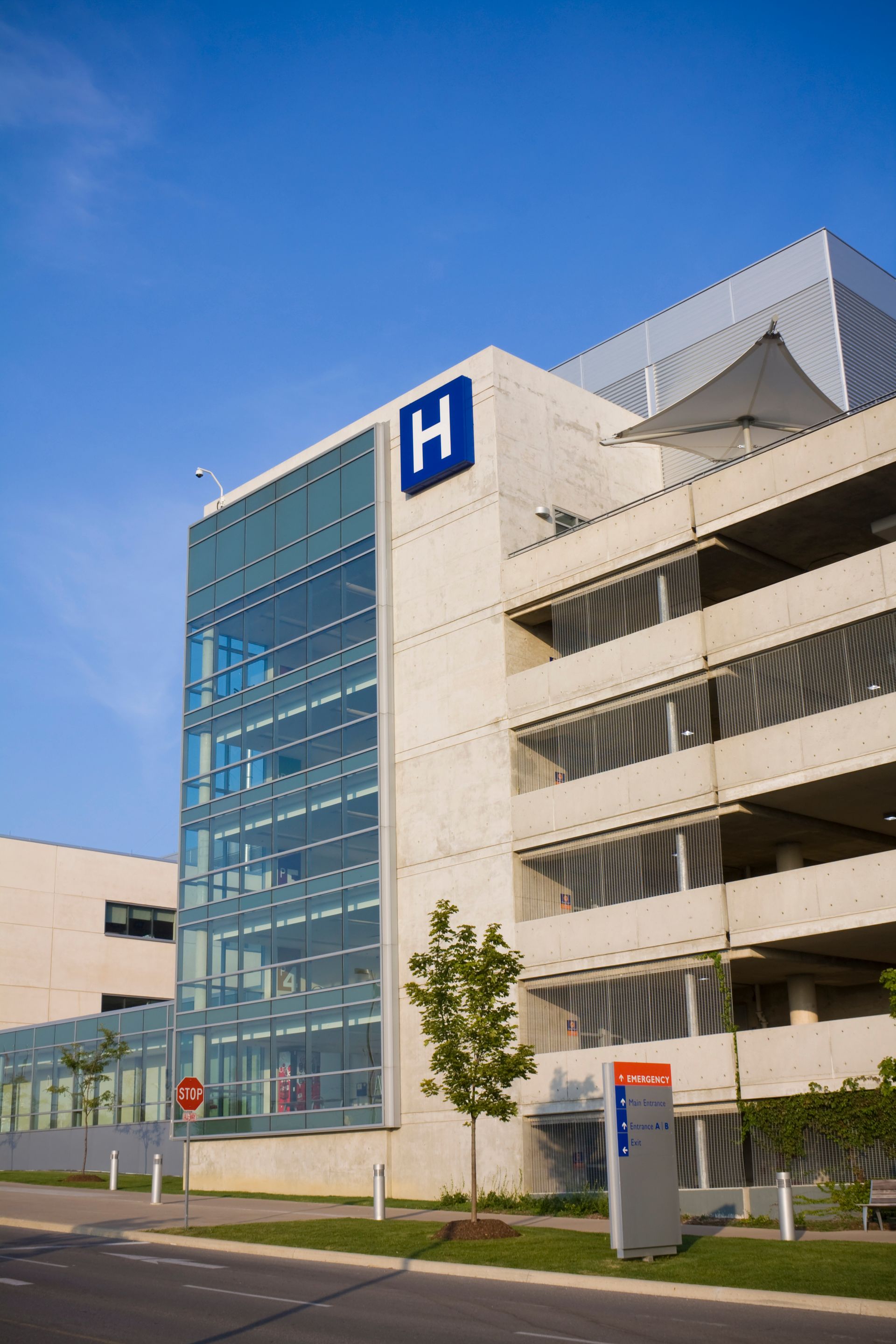 Parking moderne avec un panneau d'hôpital bleu en forme de H, une cage d'escalier vitrée et un bardage métallique.