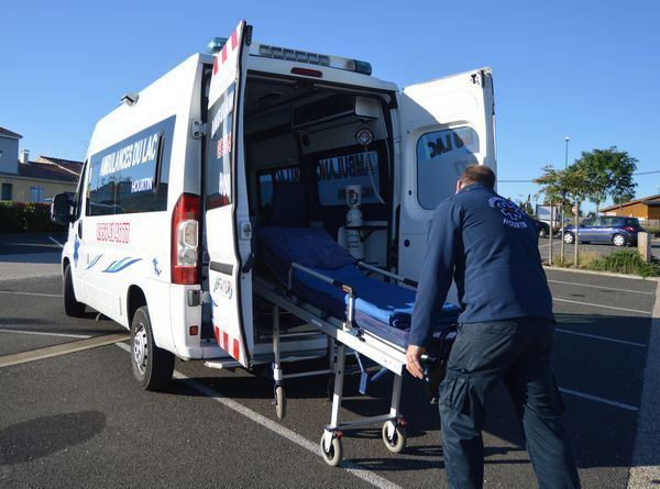 Sur un parking, une personne en uniforme sombre pousse un brancard à roulettes vers l'arrière d'une ambulance.