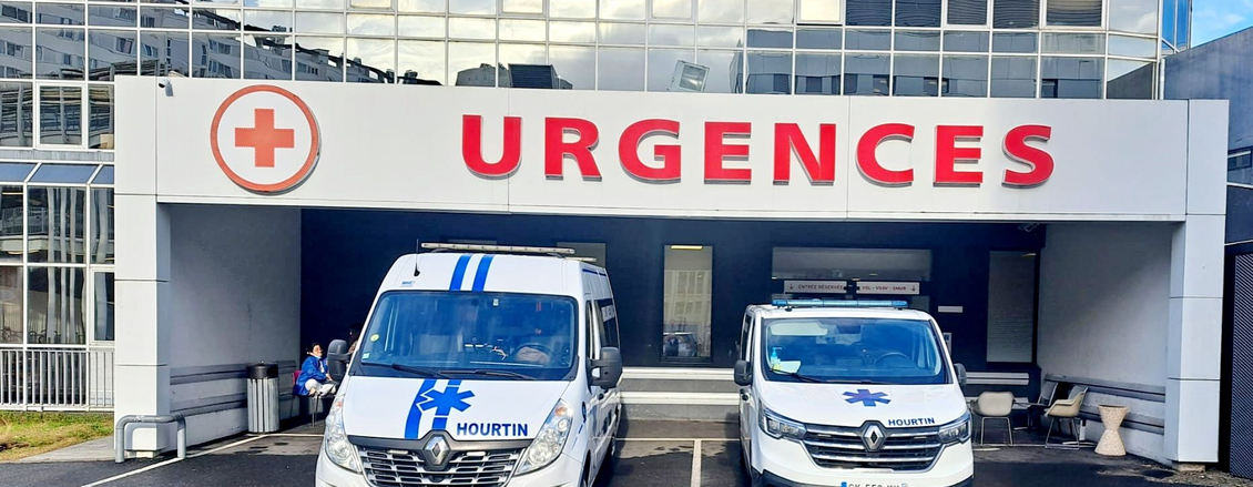 Deux ambulances stationnées sous un panneau d'entrée des urgences portant la mention URGENCES dans un hôpital.