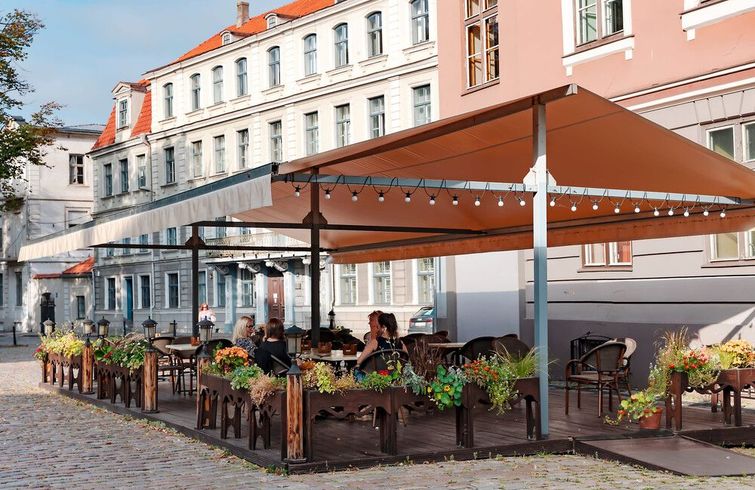 Terraza de café al aire libre con sombrillas naranjas y macetas con flores frente a edificios blancos.