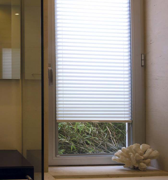 Ventana pequeña con persianas blancas, vista parcial de la vegetación exterior y un objeto decorativo blanco en el alféizar.