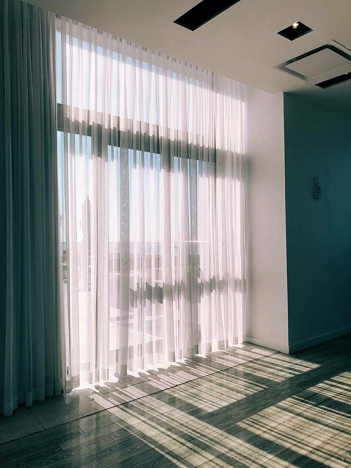 Habitación bañada por el sol, con cortinas blancas translúcidas que proyectan largas sombras sobre un suelo claro