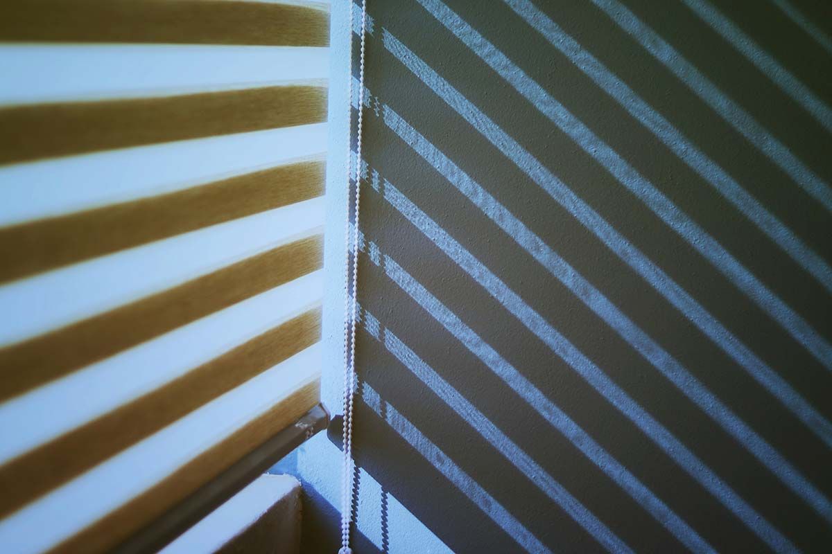 La luz del sol proyecta sombras diagonales de rayas en una pared y unas persianas en la esquina de una habitación