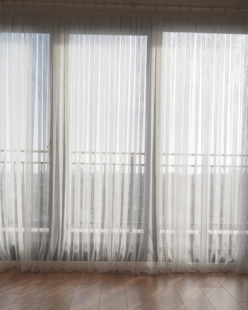 Cortinas blancas translúcidas cubren las ventanas altas de una habitación luminosa con suelo de madera.