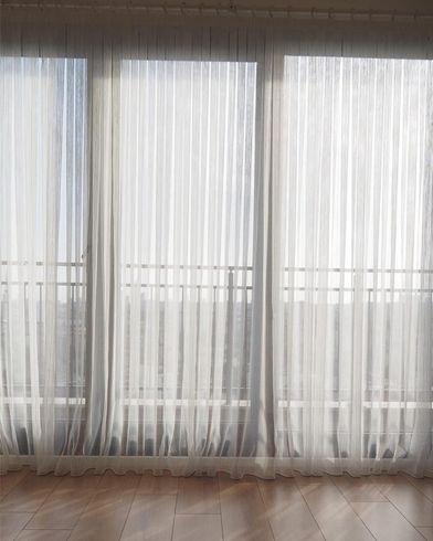 Cortinas blancas translúcidas cubren las ventanas altas de una habitación luminosa con suelo de madera.