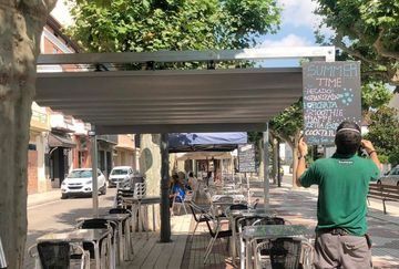 Terraza de una cafetería bajo una pérgola en una calle tranquila, con un hombre con camisa verde tomando una foto.