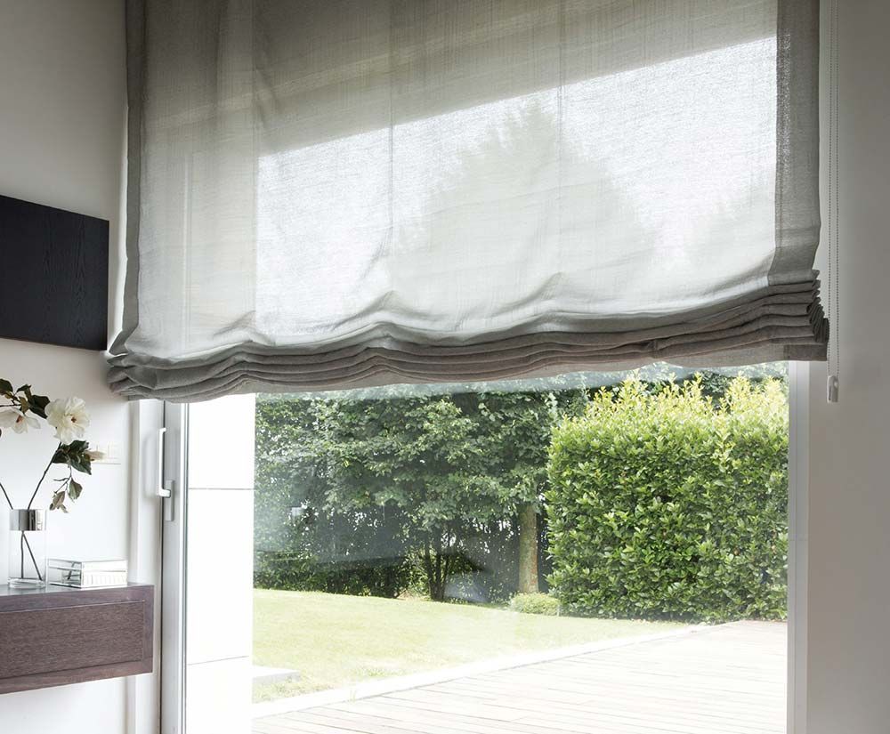 Una ventana con una persiana romana blanca parcialmente subida, con vistas a un jardín verde.
