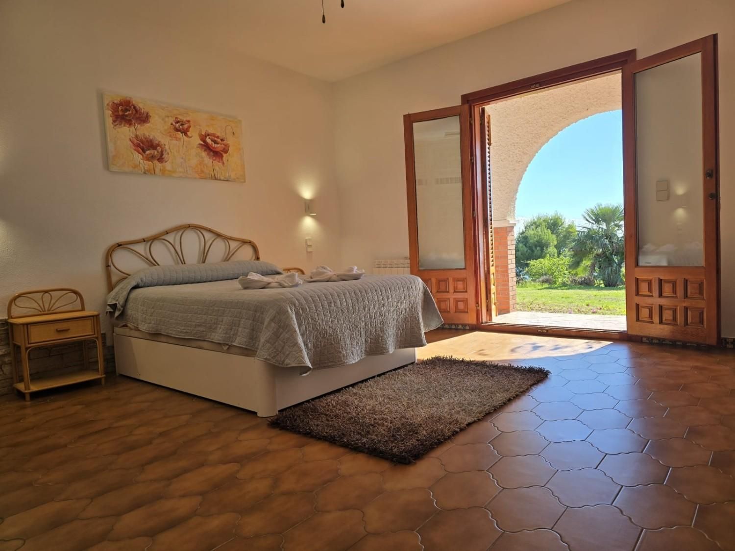 Un dormitorio luminoso con una cama, una pequeña mesita de noche y una puerta abierta que da a un jardín verde.