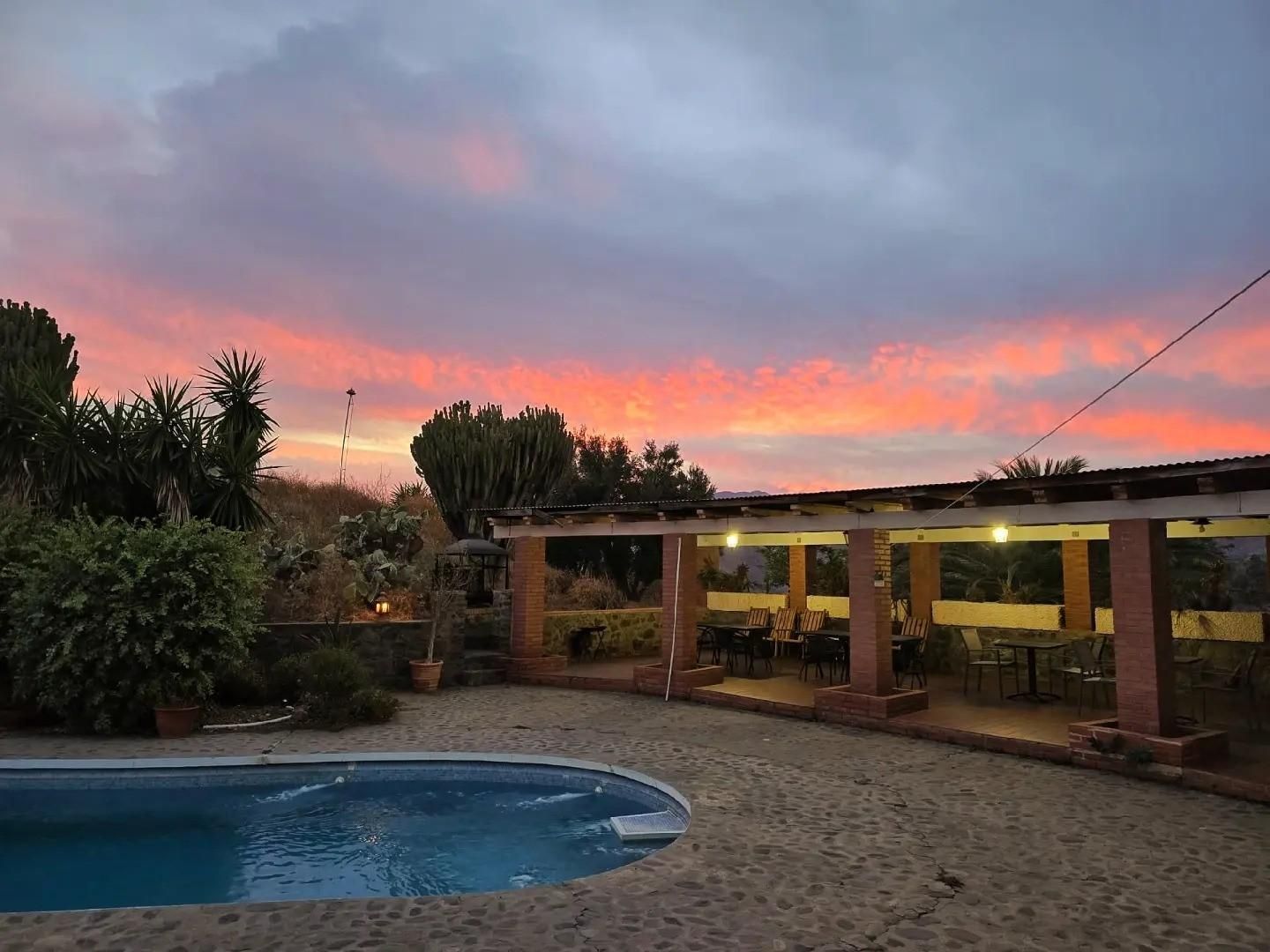 Una piscina junto a un patio con pilares de ladrillo al atardecer, con vibrantes nubes naranjas y moradas en el cielo.