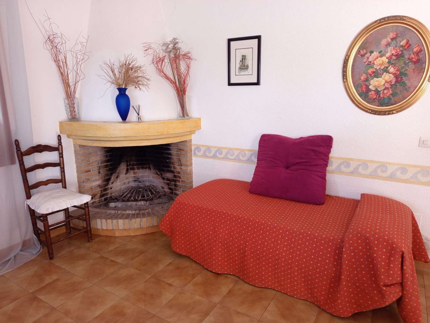 Un rincón de la habitación con chimenea, una silla y una cama individual cubierta con una colcha naranja y un cojín morado.