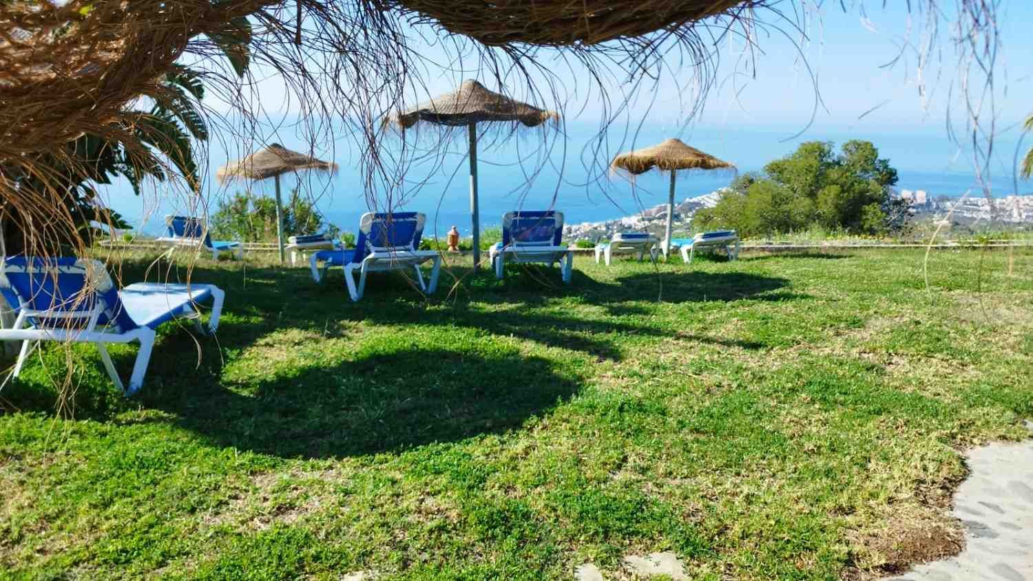 Tumbonas azules bajo sombrillas de paja en una colina cubierta de hierba con vistas a un pueblo costero y al mar.