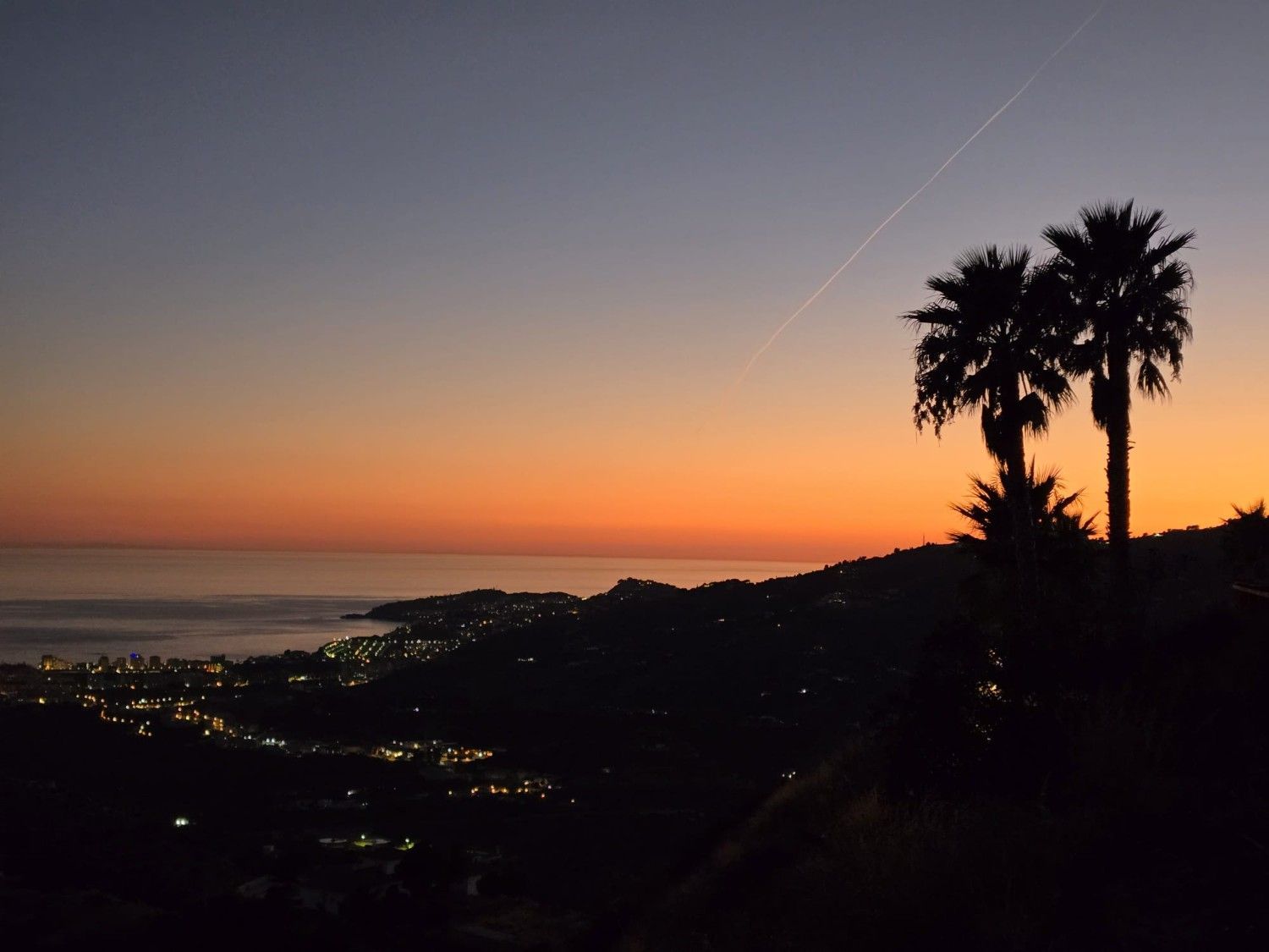 Una vibrante puesta de sol de color naranja y púrpura sobre un pueblo costero, vista tras la silueta de las palmeras.