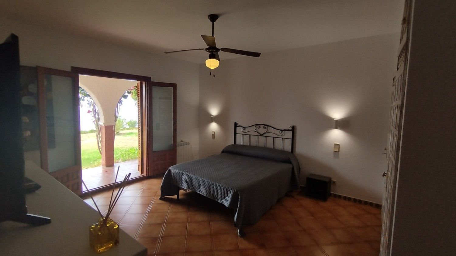 Un dormitorio con cama, ventilador de techo, lámparas de pared y puertas de cristal que dan a un espacio exterior verde.