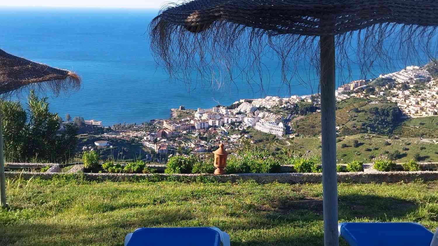 Pueblo costero visto desde una soleada ladera cubierta de hierba, con una sombrilla de paja en primer plano.