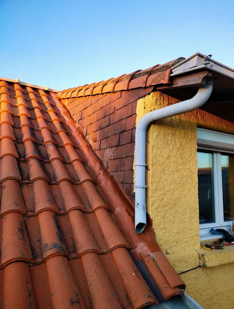 Toit de tuiles rouges avec une gouttière blanche sur un mur jaune et un ciel bleu.