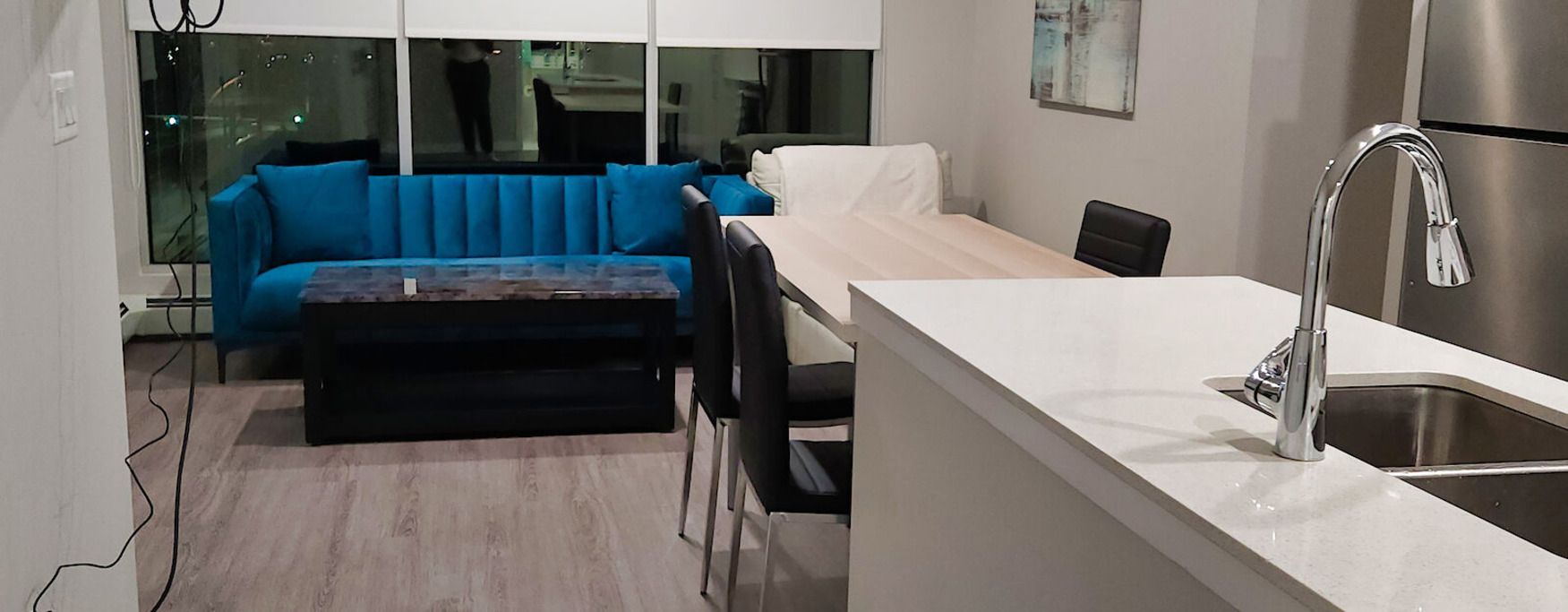 Interior of a modern apartment with a teal sofa, marble coffee table, and kitchen with stainless steel sink and faucet.
