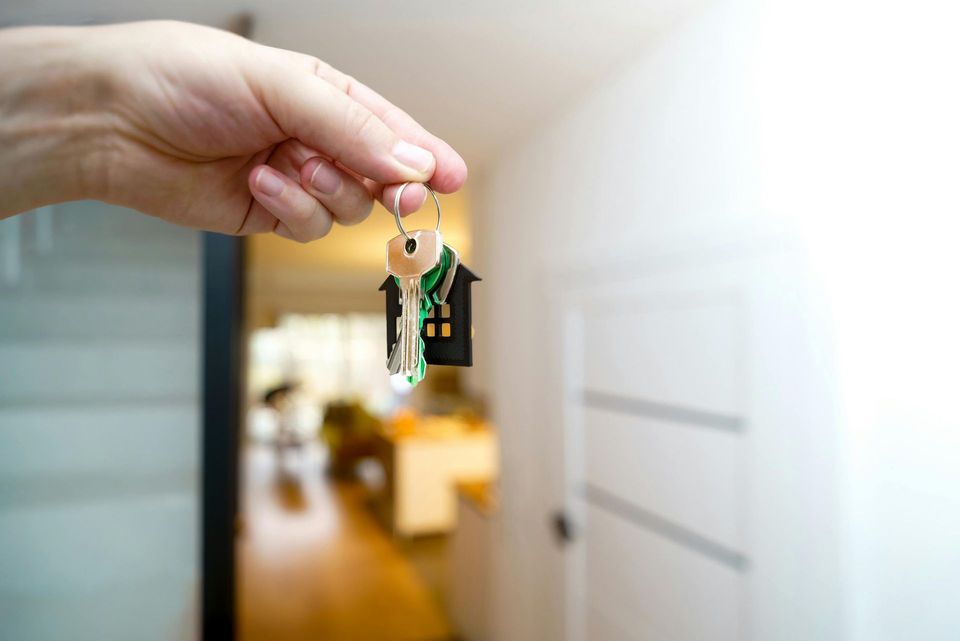 Hand holding keys with a house-shaped keychain, likely representing a new home; blurred interior in the background.