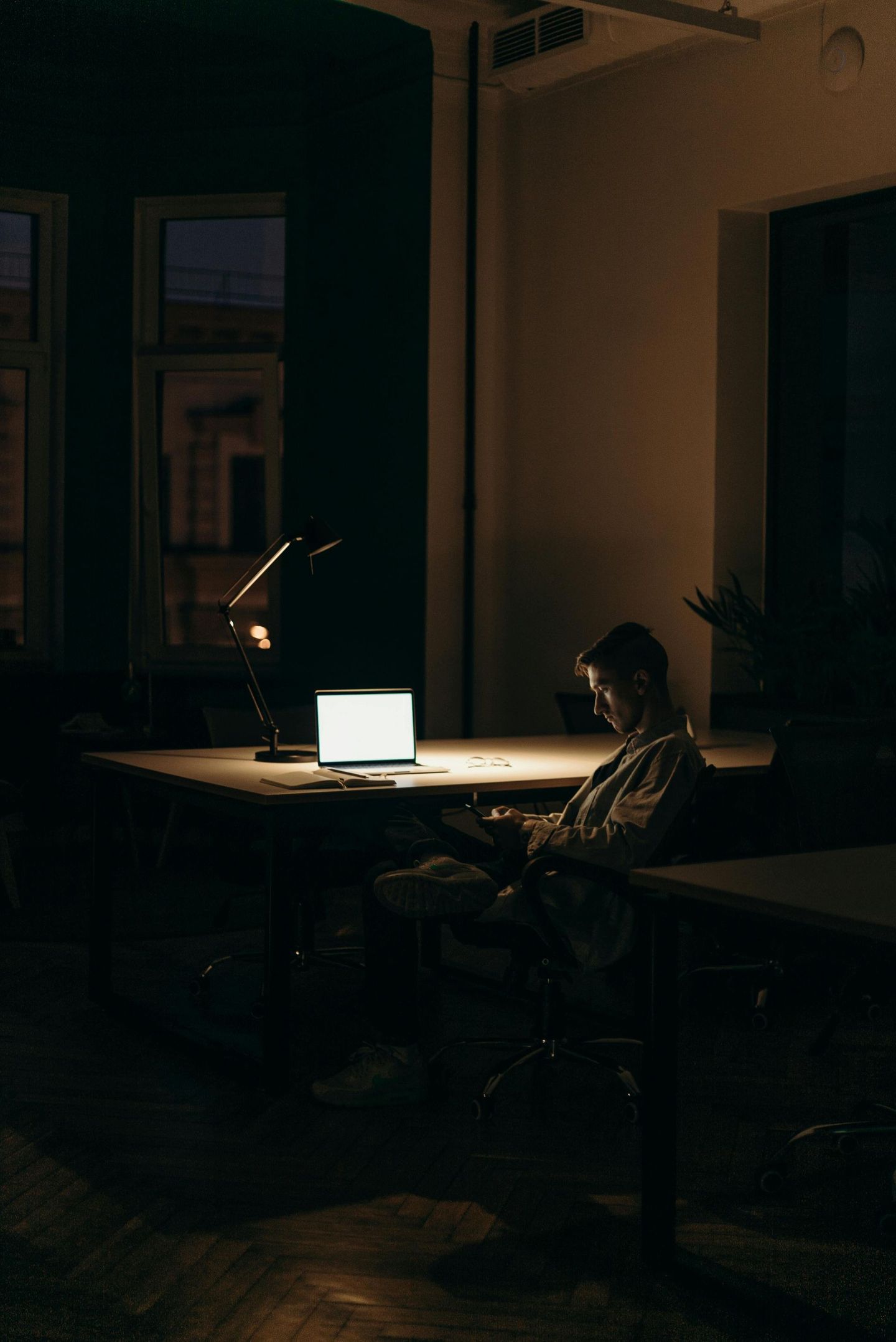 Person working on a laptop at a desk in a dimly lit room, illuminated by a desk lamp.