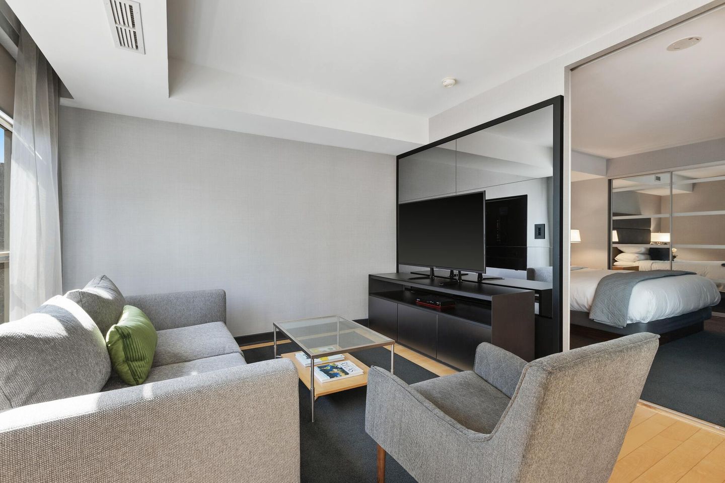Modern hotel suite with gray sofa, armchair, TV, and bedroom visible through a mirrored divider.