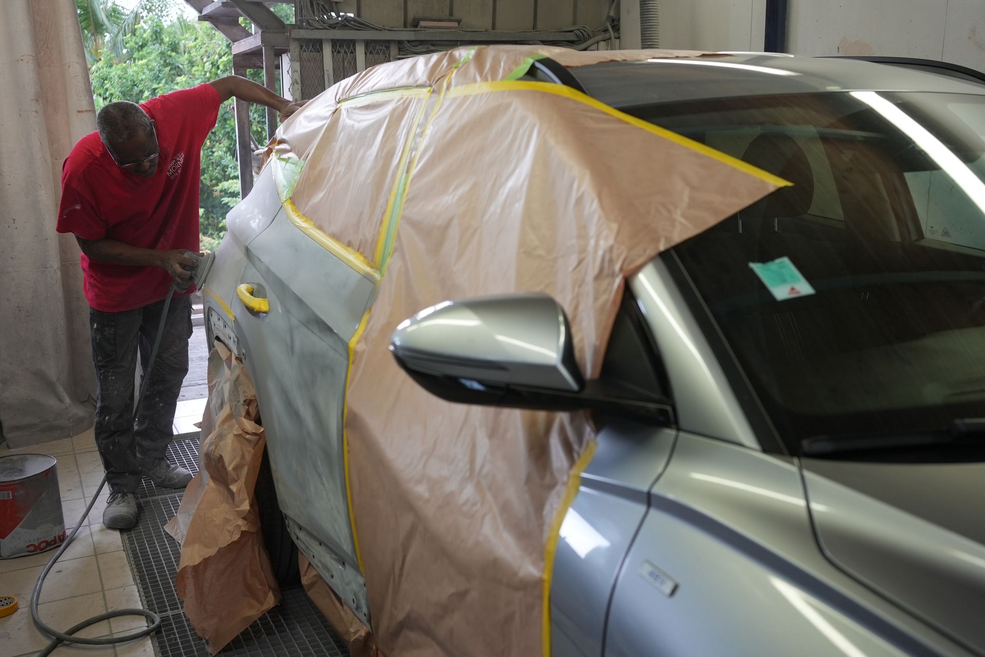 Une personne masque une voiture argentée avec du ruban de masquage et une bâche en plastique pour la peindre.