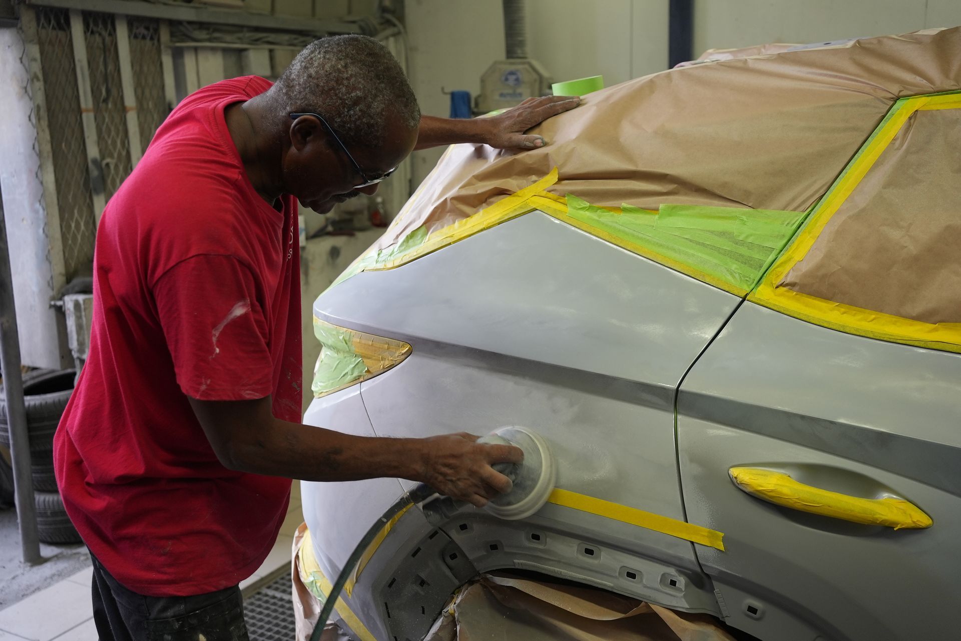 Un carrossier ponce une portière de voiture argentée, les vitres étant recouvertes de ruban adhésif et de papier.