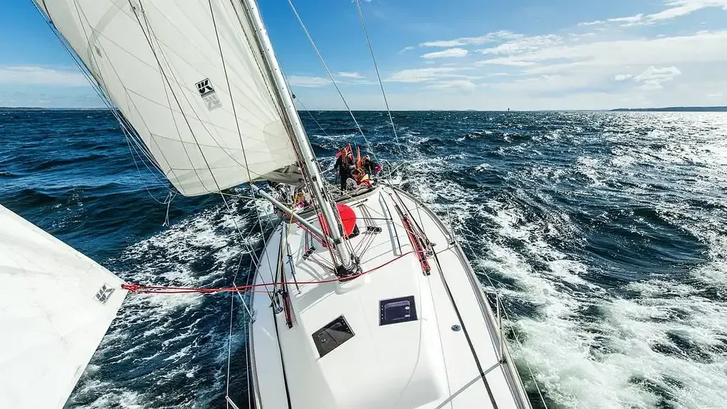 Velero en mar agitado con tripulación visible, velas blancas ondeando, agua azul.