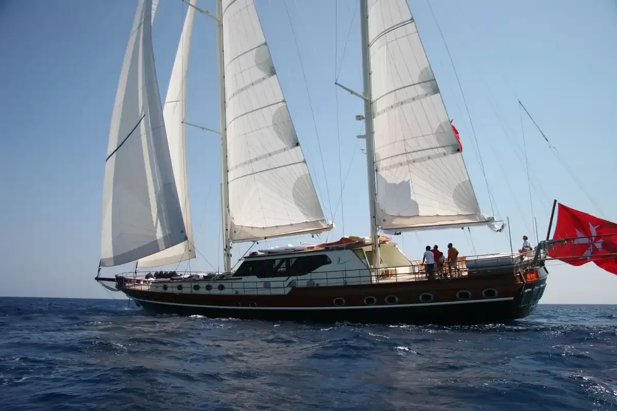 Yate de vela con velas blancas en agua azul, bandera roja en la popa y varias personas en cubierta.