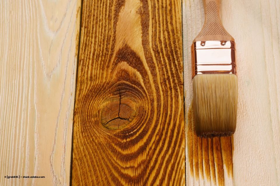 Holzplanken, eine davon braun gebeizt, mit einem Pinsel Beize aufgetragen.