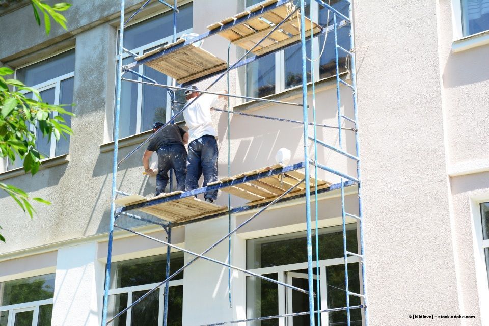 Zwei Arbeiter streichen auf einem Gerüst die Außenfassade eines Gebäudes.