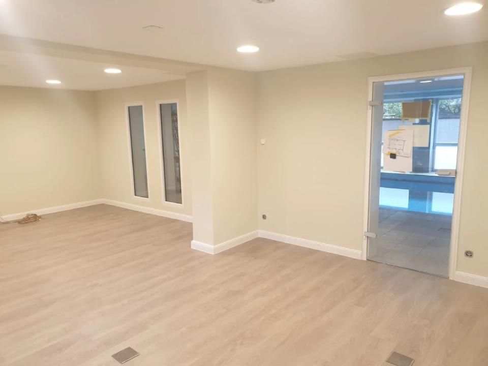 Leerer Raum mit beigen Wänden, Holzbodenoptik, Einbauleuchten und einer Tür.